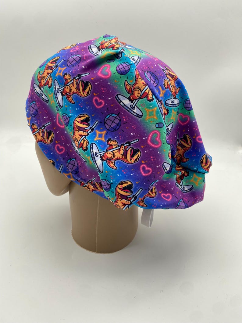 May include: A colorful scrub cap featuring a repeating pattern of cartoon dinosaurs pole dancing. The background has a vibrant gradient of purple, blue, and teal, with hearts, musical notes, and disco balls.