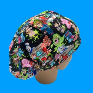 May include: A black scrub cap featuring a colorful pattern of Ghostbusters characters and the phrase "Who You Gonna Call?" The design includes images of Slimer, the Stay Puft Marshmallow Man, and other iconic figures from the movie.