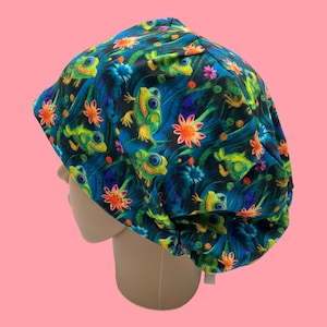 May include: A blue scrub cap featuring a whimsical pattern of green and yellow frogs, orange and pink flowers, and blue water-like lines. The cap is designed to cover the head and hair, commonly used in medical or culinary settings.