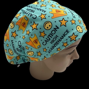 May include: A teal surgical scrub cap with a repeating pattern of yellow stars, smiley faces, and caution signs. The words "CAUTION HIGH MAINTENANCE" are printed on the cap in navy blue. The cap is designed to cover the head.