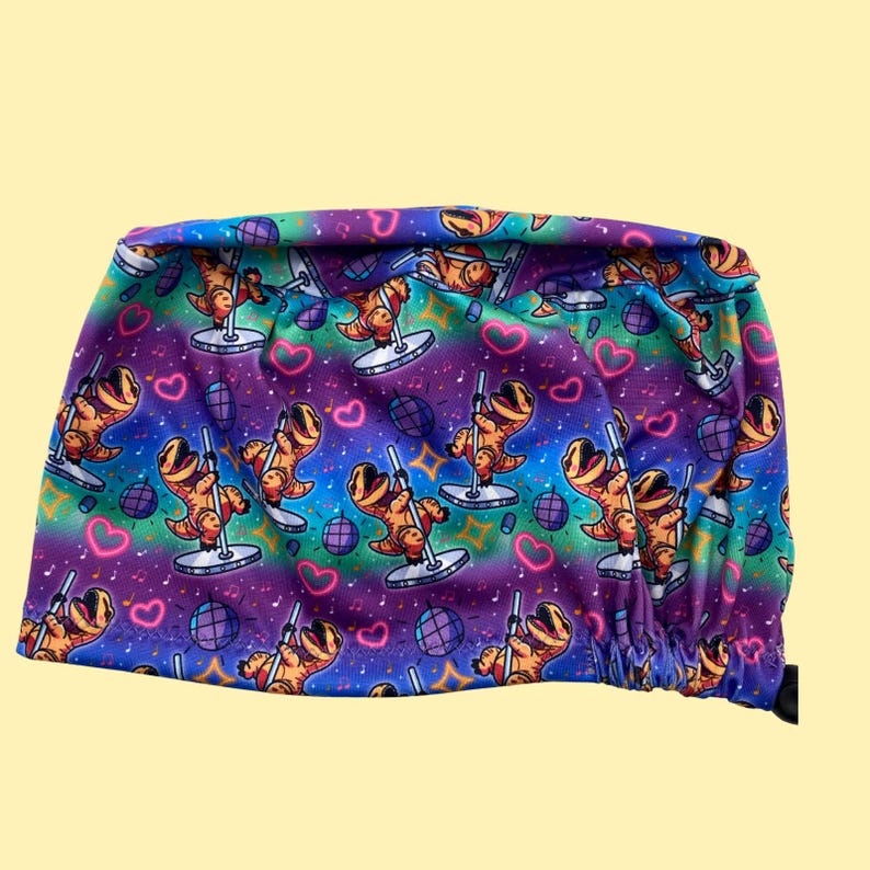 May include: A colorful scrub cap featuring a repeating pattern of cartoon dinosaurs pole dancing. The design includes hearts, musical notes, and disco balls on a gradient background of blue, purple, and green.