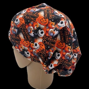 Może przedstawiać: Czapka chirurgiczna o tematyce Halloween z powtarzającym się wzorem pomarańczowych dyń, pluszowych misiów i postaci podobnych do Jacka Skellingtona na czarnym tle. Fraza "Scaring is Caring" jest wydrukowana.