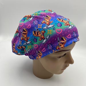 May include: A colorful scrub cap featuring a repeating pattern of orange cartoon dinosaurs pole dancing. The background is a vibrant mix of purple, blue, and green with hearts, disco balls, and musical notes.