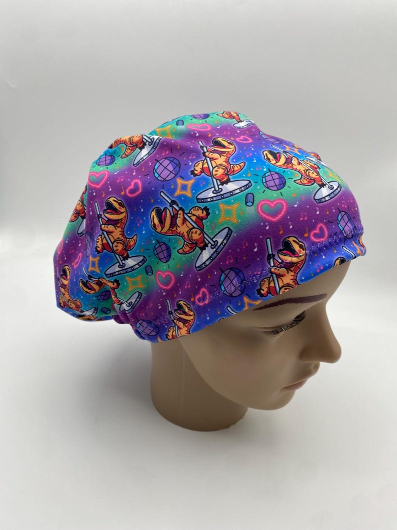 May include: A colorful scrub cap featuring a repeating pattern of orange cartoon dinosaurs pole dancing. The background is a vibrant mix of purple, blue, and green with hearts, disco balls, and musical notes.