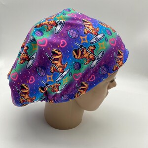 May include: A colorful scrub cap featuring a repeating pattern of cartoon dinosaurs pole dancing. The background has a rainbow gradient with hearts, disco balls, and stars. The cap is designed to fit comfortably and securely.