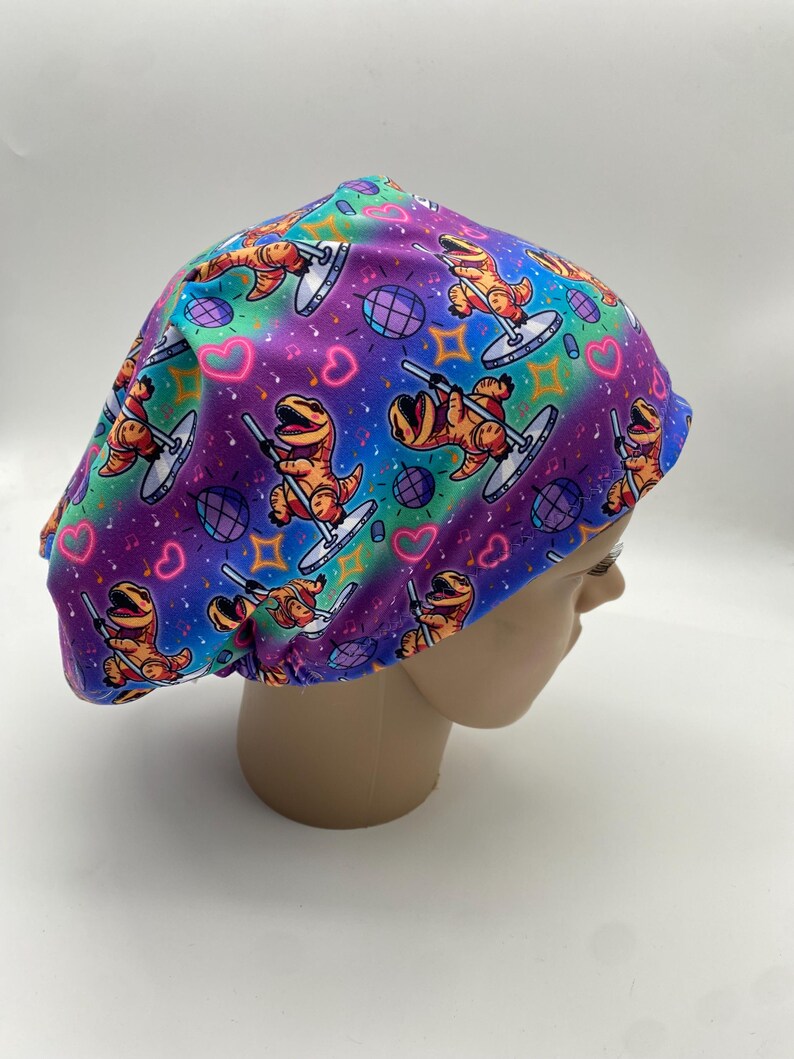 May include: A colorful scrub cap featuring a repeating pattern of cartoon dinosaurs pole dancing. The background has a rainbow gradient with hearts, disco balls, and stars. The cap is designed to fit comfortably and securely.