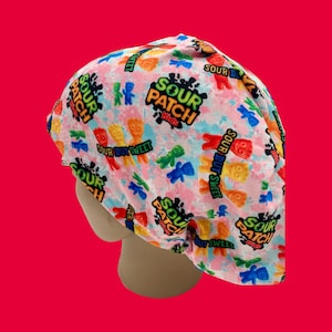 Op de afbeelding: Een roze chirurgische scrub-muts met een herhalend patroon van Sour Patch Kids snoepjes, met het Sour Patch Kids logo en de zin "Sour But Sweet".