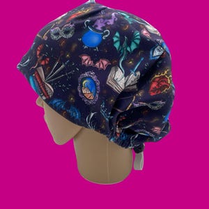 Puede incluir: Un gorro quirúrgico azul oscuro con un patrón repetido de elementos fantásticos. El diseño incluye dragones, calderos de pociones, murciélagos, libros y otras ilustraciones temáticas de fantasía. El gorro está diseñado para adaptarse a la cabeza y tiene una corbata blanca.
