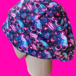 May include: A scrub cap featuring a vibrant pattern of skulls and flowers in shades of neon pink, electric blue, and purple against a black background. The fabric has a gathered design with a rounded crown and a structured fit for head coverage.