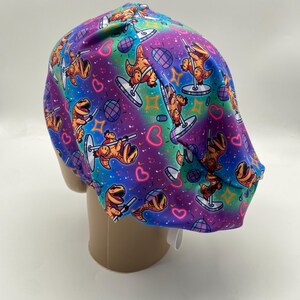 May include: A colorful scrub cap featuring a repeating pattern of orange dinosaurs pole dancing on a purple, blue, and green background. The design includes hearts, disco balls, and stars. The cap is made of a soft, flexible material.
