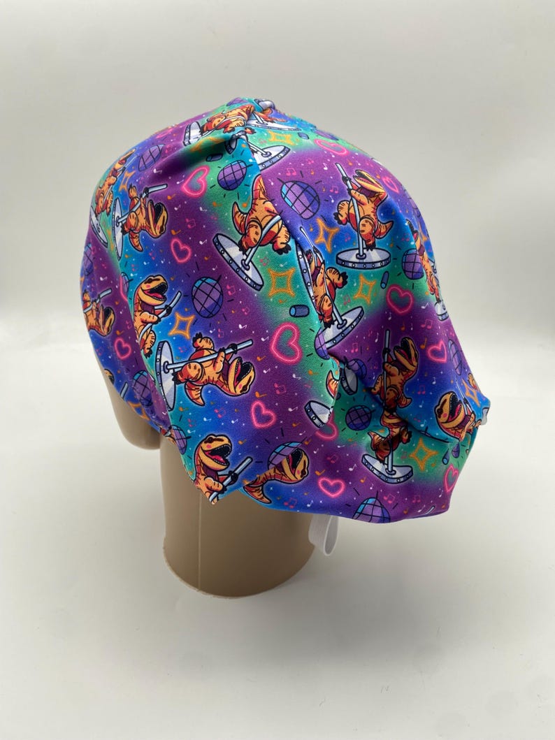 May include: A colorful scrub cap featuring a repeating pattern of orange dinosaurs pole dancing on a purple, blue, and green background. The design includes hearts, disco balls, and stars. The cap is made of a soft, flexible material.