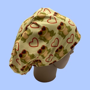 Puede incluir: Un gorro quirúrgico amarillo claro con un patrón repetido de ranas verdes con gafas de sol rojas en forma de corazón y contornos de corazón rojos. El gorro está hecho de una tela suave y está diseñado para un ajuste cómodo.