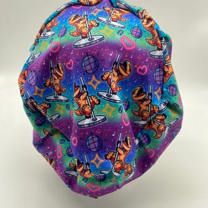 May include: A colorful scrub cap featuring a repeating pattern of orange Tyrannosaurus Rex dinosaurs pole dancing. The background transitions from teal to purple, with disco balls, hearts, and musical notes.