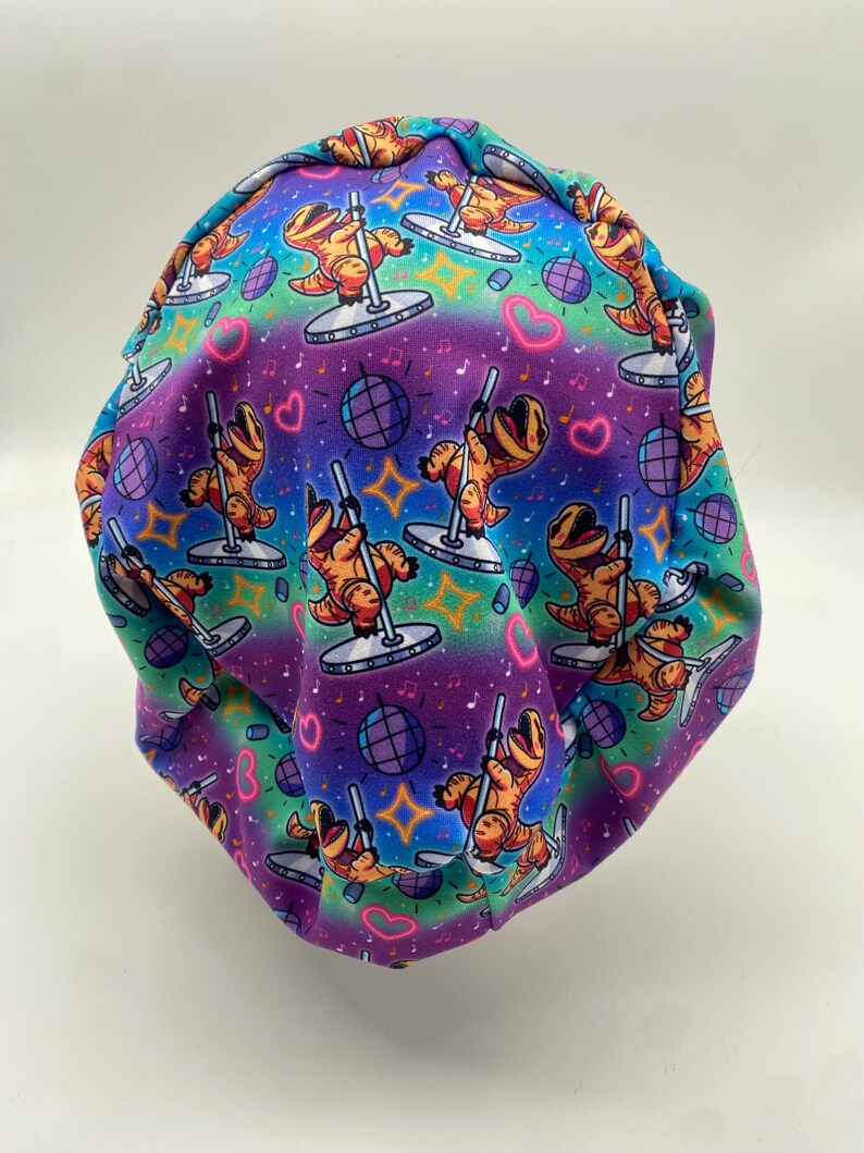 May include: A colorful scrub cap featuring a repeating pattern of orange Tyrannosaurus Rex dinosaurs pole dancing. The background transitions from teal to purple, with disco balls, hearts, and musical notes.