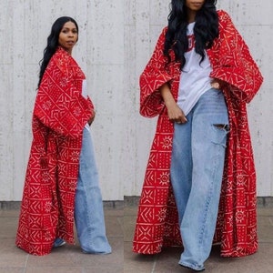 Op de afbeelding: Twee vrouwen dragen lange, rood-witte gepatroneerde gewaden met lange mouwen. De gewaden hebben een geometrisch patroon met witte zonnen en lijnen op een rode achtergrond. Beide vrouwen dragen een blauwe jeans.