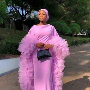 May include: A woman wearing a long, light purple dress with ruffled sleeves and a matching headscarf. She is holding a small black purse.