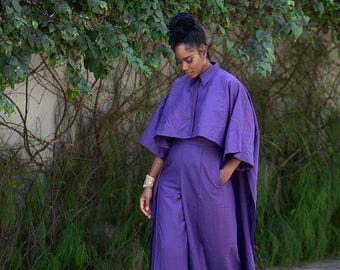 May include: A woman wearing a purple jumpsuit with a cape-like top. The jumpsuit has a collared shirt style top and a wide leg silhouette.