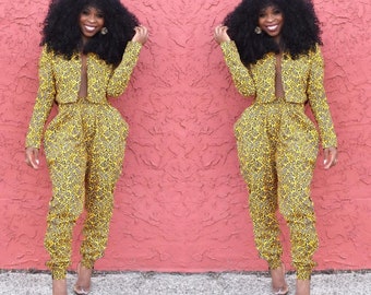 May include: A woman is wearing a yellow and black patterned pantsuit with a cropped jacket. The pantsuit has a loose fit and the jacket has a V-neckline. The woman is standing in front of a pink wall.