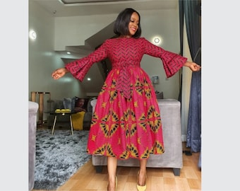 May include: A woman wearing a red and black patterned dress with long sleeves and a belt. The dress has a flared skirt and a geometric pattern. She is standing in a room with a gray rug and a couch in the background.