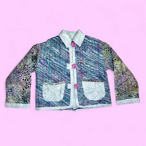May include: A quilted jacket featuring a blue and purple wavy pattern with contrasting white floral print pockets, collar, and cuffs. The garment includes three bright pink buttons down the center front, long sleeves, and a structured collar design.