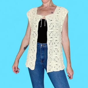 May include: Cream-colored crochet vest with a square neckline and open front. The vest features a floral pattern and a tie closure at the bust. The model is wearing a black top and blue jeans.