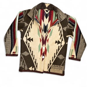 May include: A long-sleeve button-up jacket featuring a cream-colored center panel with a dark brown geometric diamond pattern. The sleeves and sides display multi-colored stripes in red, teal, and black. A small rectangular label reads Spirit of the Peoples.