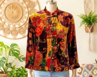 May include: A long-sleeve velvet jacket featuring a vibrant floral pattern in shades of yellow, red, purple, and green. The jacket has a mandarin collar, three-quarter length sleeves, and a front closure secured with five decorative fabric toggle buttons.