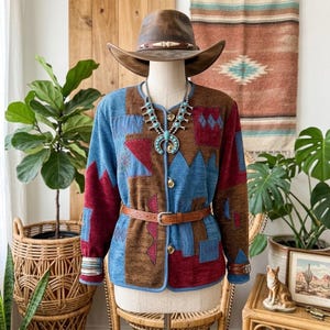 May include: A long-sleeved jacket featuring a patchwork design in shades of blue, brown, and red, accented with a turquoise squash blossom necklace, a brown leather belt, and a brown cowboy hat, all displayed on a mannequin.