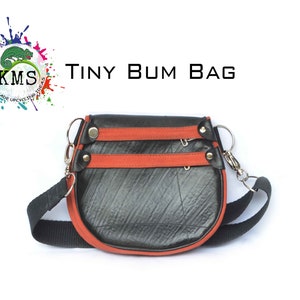 May include: A small black and orange bum bag made from recycled tire inner tubes. The bag has two zippered compartments and a black adjustable strap. The bag is labeled "TINY BUM BAG".