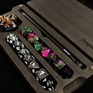 May include: A dark wooden rectangular case with the text Dungeons Box engraved on the lid. The interior features custom slots holding a set of black dice with white numbers, a set of iridescent multi-coloured dice, a small figurine, and a black pencil.