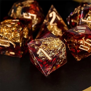 May include: A set of polyhedral dice made from translucent deep red resin with embedded gold leaf flakes. Each die features sharp edges and clear, recessed numbers filled with a metallic gold finish, creating a high-contrast appearance.