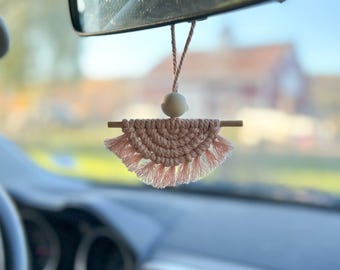 May include: A pink macrame car air freshener hanging from a rearview mirror. The air freshener is shaped like a half-circle with fringe on the bottom and a wooden dowel across the top. A small white bead hangs from the top of the air freshener.