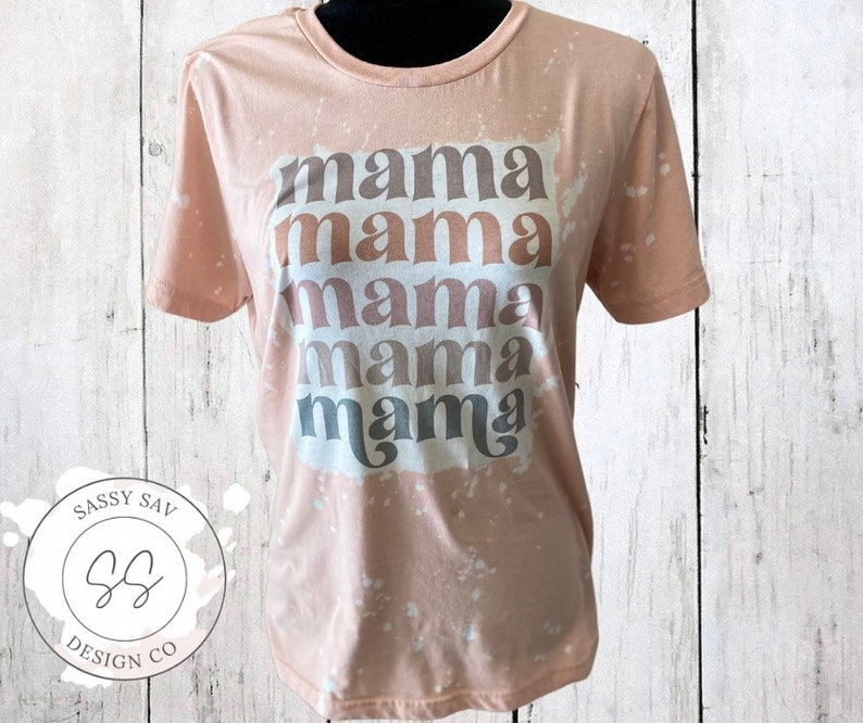 May include: A light pink t-shirt with a bleach-splattered design. The shirt has the word "mama" repeated five times in a stacked design, with each word in a different shade of brown.