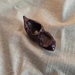 Könnte beinhalten: Ein kleines, dunkelviolettes Keramikobjekt in Schuhform mit glänzender Oberfläche. Das Stück hat eine spitze Zehenpartie und eine offene Oberseite, die an einen Miniatur-Clog erinnert. Die Oberfläche weist subtile Farb- und Texturvariationen auf.