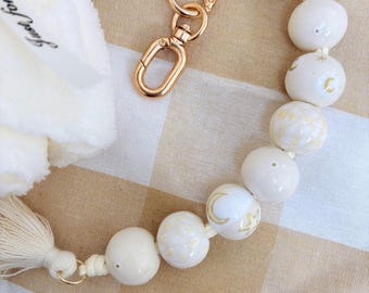 Peut inclure: Un porte-clés composé de perles en céramique crème lisse ornées de motifs de lunes et d'étoiles dorées. Les perles sont enfilées sur un cordon avec un fermoir mousqueton en métal doré à une extrémité et un pompon en tissu crème à l'autre.