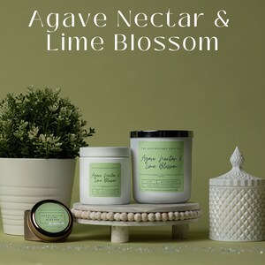 May include: Three candles in white jars with silver lids. The candles are labeled "Agave Nectar & Lime Blossom". The candles are on a white marble surface with a wooden bead trim. The background is a light green.