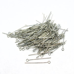 May include: A pile of silver-toned head pins with eye loops. The head pins are scattered on a white surface.