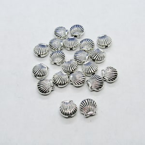 May include: A collection of silver-toned metal seashell-shaped beads. The beads are arranged in a pile on a white surface.