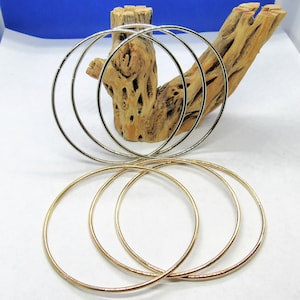 May include: Four silver and four gold bangle bracelets. The silver bracelets are stacked on a piece of driftwood. The gold bracelets are stacked on a white surface.