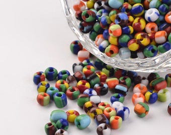 May include: A bowl filled with colorful glass seed beads. The beads are a mix of red, blue, green, yellow, and white. The beads are scattered on a white surface.