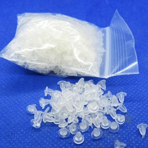 May include: A clear plastic bag filled with small, clear plastic earring backs. The bag is open and the earring backs are spilling out onto a blue surface.