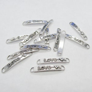 May include: A group of silver-toned metal charms with the word "Love" and a heart design. The charms are shaped like a rectangle with rounded edges.