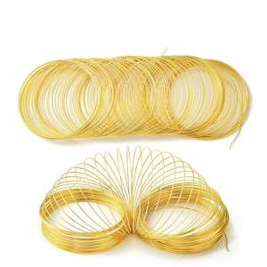 May include: Several coils of gold-colored wire are arranged on a white background. The wire is thin and appears to be made of metal. The coils are circular and neatly stacked, suggesting a craft or jewelry-making supply.