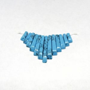 May include: A strand of turquoise blue gemstone beads, each bead is a long, thin, rectangular shape with a slightly rounded edge. The beads are arranged in a fan shape, with the longest beads at the top and the shortest beads at the bottom.