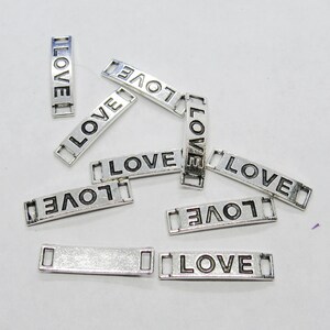May include: A group of silver-toned metal charms with the word "LOVE" engraved on them. The charms are rectangular and have small holes at each end for attaching to a chain or bracelet.
