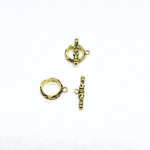May include: Gold-toned metal toggle clasp with an ornate design. The clasp is made of two pieces: a round ring and a bar with a loop at each end.