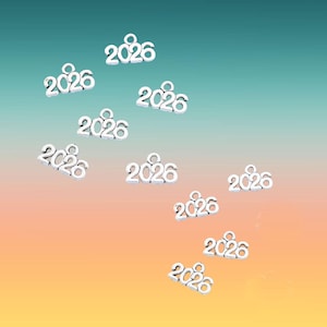 May include: Scattered silver-colored charms, each displaying the year "2026". The charms have a small loop at the top for attaching to jewelry or other crafts. The background transitions from blue to orange.