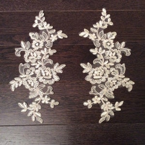 May include: Two ivory floral lace appliques. Each applique features a detailed design of flowers and leaves, with a delicate, openwork pattern. The appliques are set against a dark brown wooden surface.