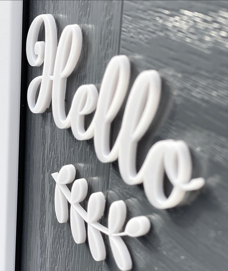 May include: White 3D letters spelling "hello" with a white 3D leaf branch underneath. The letters and branch are attached to a dark grey surface.