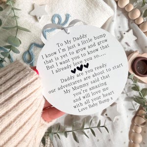 May include: A white, round baby announcement with the text "To My Daddy" and a loving message from "Love Baby Bump x." The announcement is held by a person wearing a beige sweater. Decorative stars and greenery surround the announcement.